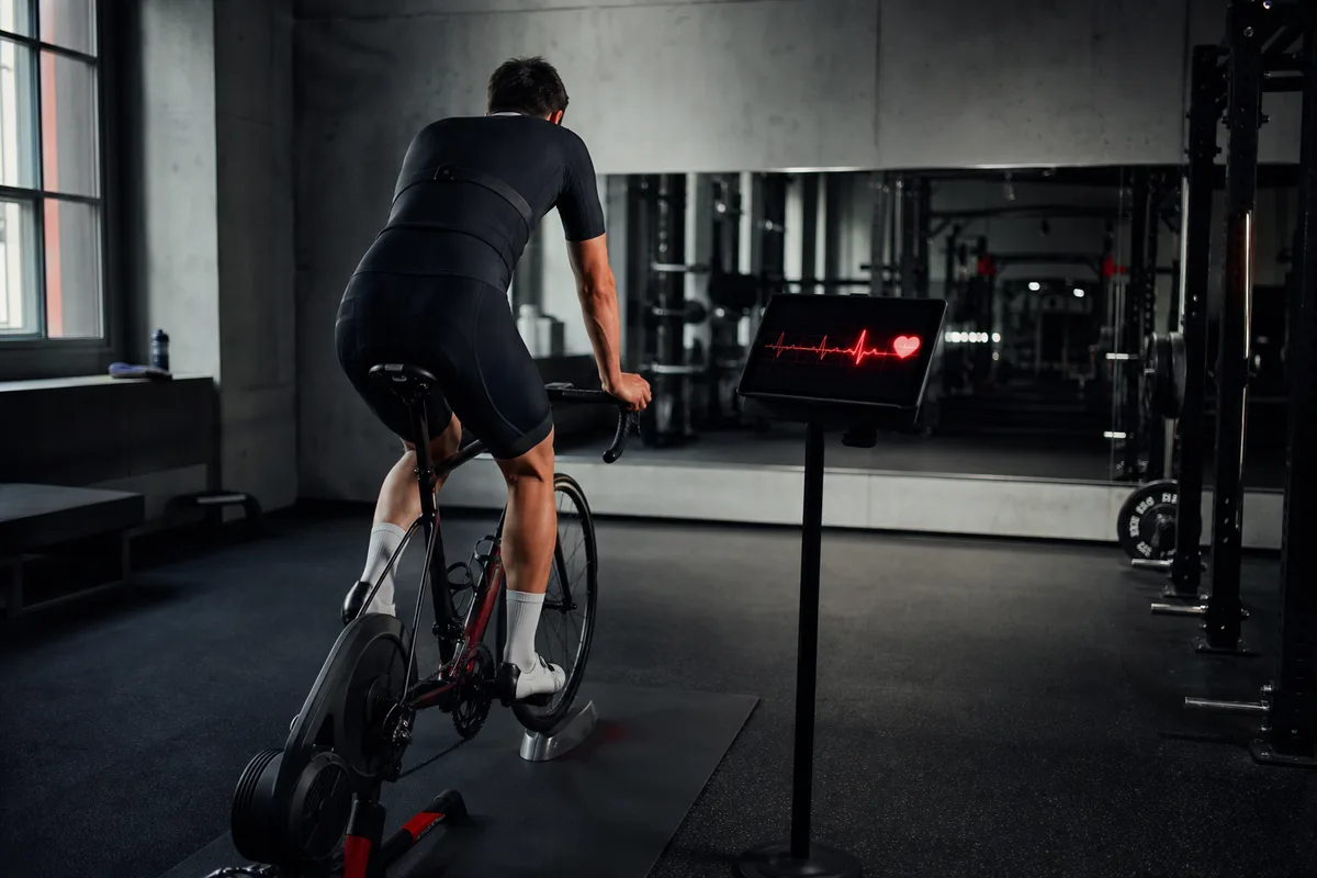 Cycliste sur home-trainer en salle avec ceinture cardiofréquencemètre et écran affichant les pulsations